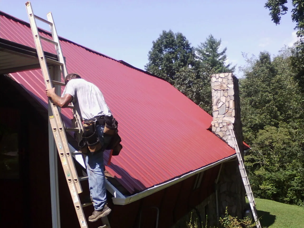 Roofer on a ladder beside a red metal roof and stone chimney — Soffit & Fascia Repair by Roofing Brothers in Millport